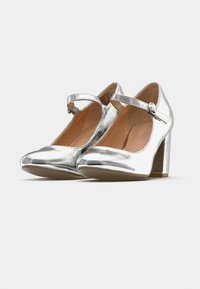 Zapatos de tacón Mary Jane metálicos plateados con punta redonda, correa en el tobillo y un tacón bloque de altura media. Textura suave con un acabado pulido.