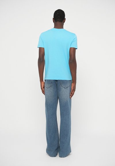 Turkizna bombažna majica s kratkimi rokavi in okroglo ovratnikom, v kombinaciji z širokimi svetlo modrimi jeans hlačami, gledano s hrbta.