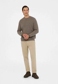 Maglione grigio a costine, pantaloni beige e scarpe marroni indossati da un uomo in piedi. Il maglione ha una scollatura rotonda e maniche lunghe.