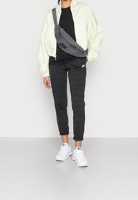 Ljusgrön zip-jacka, svart t-shirt, grå slingväska, mörka mönstrade joggers och vita sneakers. Slät tyg, avslappnad passform, logotypdetaljer.