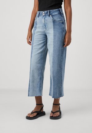 Frau trägt zweifarbige, weite, cropped Jeans und schwarze Sandalen mit Knöchelriemen, steht vor einem schlichten hellen Hintergrund.