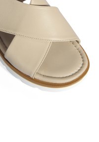 Beige leren sandaal met een kruisbandontwerp, open teen en een gestructureerde voetbed. Voorzien van een rubberen zool met een contrasterende bruine rand.