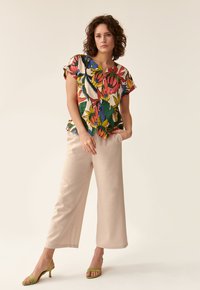 Bluză cu model floral, cu mâneci scurte, design multicolor, cu nasturi în față; asortată cu pantaloni bej largi și sandale verzi.