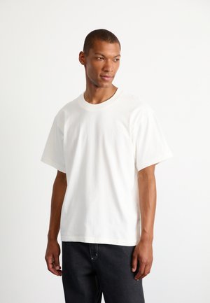 Hvid bomulds T-shirt med rund hals og korte ærmer. Har et diskret logo på brystet. Parret med mørke jeans.