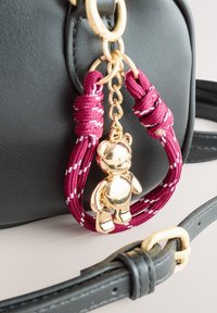 Zwarte leren handtas met een gouden teddybeerbedel bevestigd aan een bordeauxrood-wit koord, met gouden metalen accenten en een strak ontwerp.