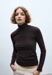 Brązowa bluzka z długim rękawem i golfem, wykonana z miękkiego materiału, o dopasowanym kroju, zestawiona z jasnymi dżinsami.