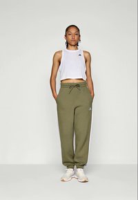 Top cropped branco com logotipo preto, calças de treino verde-oliva com riscas brancas laterais e ténis cinzentos. Design atlético simples.