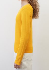Femme aux cheveux bouclés portant un pull jaune à manches longues texturé et un pantalon blanc, vue de profil sur un fond uni.