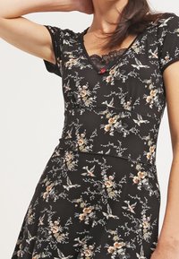 Svart blommig klänning med korta ärmar, som har V-ringning och spetsdetalj vid halsringningen. Mönstret innehåller blommor och fåglar. Bekväm passform.