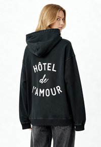 Fekete kapucnis pulóver laza szabással, a hátsó részén nagy, fehér szöveges nyomattal, ami a "HÔTEL de L'AMOUR" feliratot tartalmazza. Puha, pamut anyag, textúrált felület.