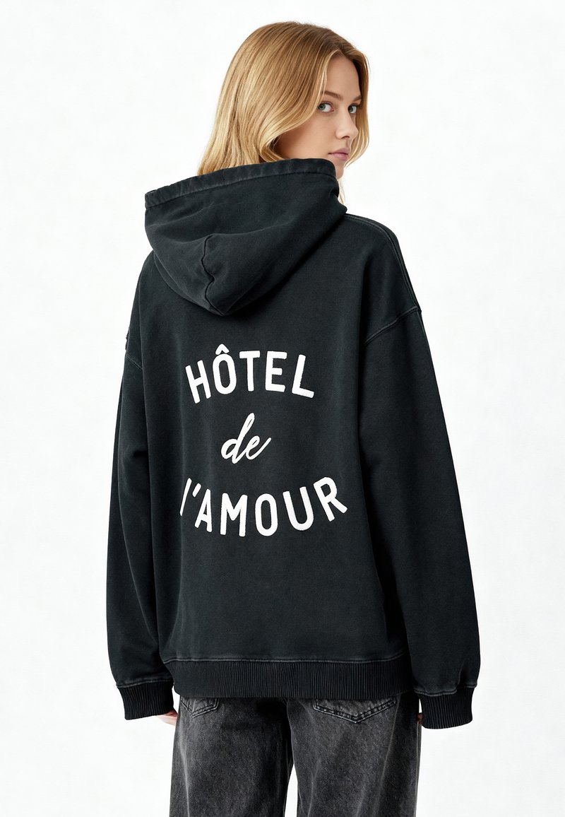 Fekete kapucnis pulóver laza szabással, a hátsó részén nagy, fehér szöveges nyomattal, ami a "HÔTEL de L'AMOUR" feliratot tartalmazza. Puha, pamut anyag, textúrált felület.