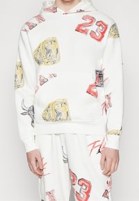 Sudadera blanca con impresiones coloridas en estilo de boceto de logotipos y números, con un bolsillo frontal y puños acanalados para un ajuste ceñido.
