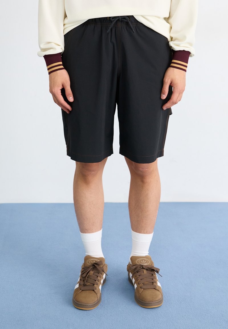 Shorts noirs jusqu'aux genoux avec taille à cordon de serrage, présentant des surpiqûres brunes et des poches latérales. Associés à des baskets brunes et des chaussettes blanches.