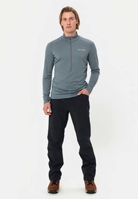 Grå långärmad topp med kvartszip och Vaude-logotyp, kombinerad med svarta byxor och bruna vandringskängor, stående mot en vit bakgrund.