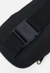 Svart tygpåse med en texturerad yta, som har en hållbar plastspänne med en logotyp och ett zip-fack längst ner.