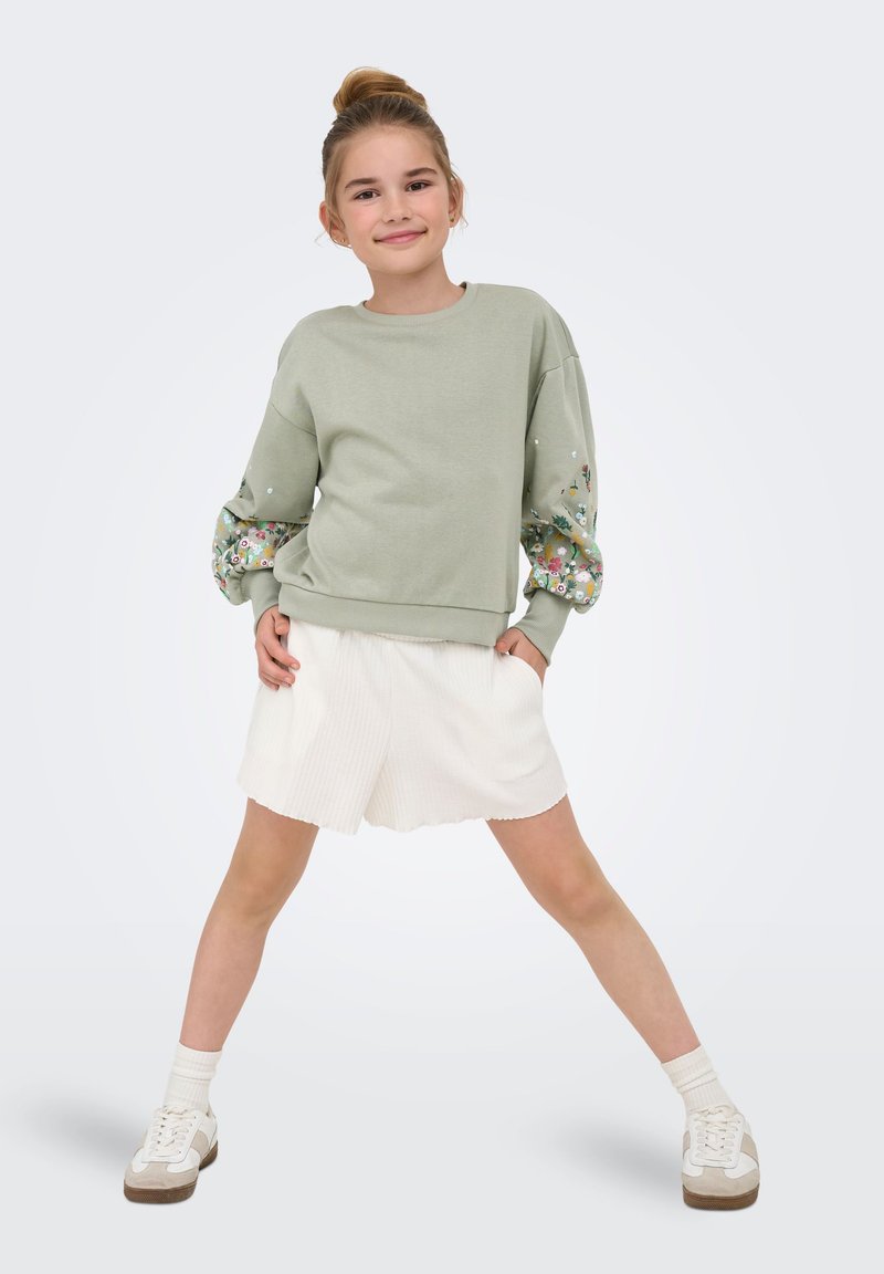 Fille debout, jambes écartées, mains dans les poches, portant un sweat-shirt vert clair avec des manches fleuries et un short blanc sur un fond uni.