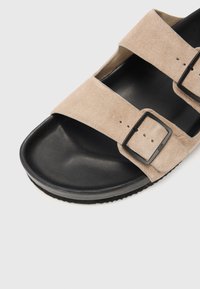 Suede sandaler med to brede beige stropper med sorte rektangulære spænder, en formet sort fodseng og en tekstureret ydersål for godt greb.