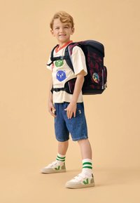 Mochila con un patrón rojo oscuro, camiseta de niño con estampados de animales multicolores, pantalones cortos de mezclilla, calcetines a rayas verdes y zapatillas blancas.