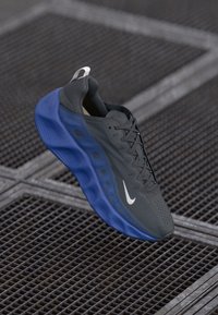Shoe desportivo da Nike com parte superior preta, sola azul, tecido texturizado, detalhes perfurados e logotipo branco de lado, exibido sobre uma grelha metálica.