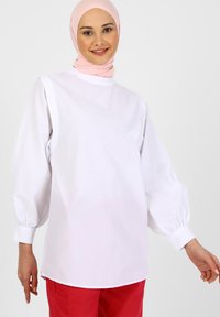 Weißes langärmeliges Oberteil mit hohem Kragen, Puffärmeln und gesäumten Säumen, kombiniert mit roten Hosen. Glattes Baumwollmaterial ohne Muster.