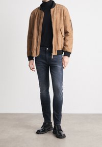 Blouson aviateur en daim beige avec fermeture éclair à l'avant, poignets côtelés, col roulé noir, jean foncé et chaussures noires à lacets avec semelles épaisses.