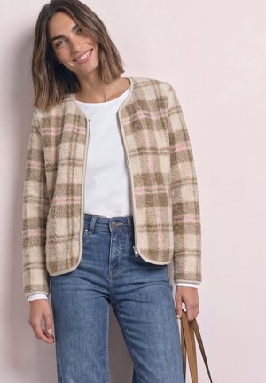 Kvinde iført beige og lyserød ternet lynlåsjakke, hvid skjorte, blå jeans og holder en brun håndtaskerem, smilende mod en lys baggrund.