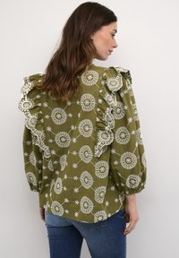 Olivgrön blus med vita cirkulära mönster och intrikat spetsdetalj på volangiga axlar. Långa ärmar med rynkade muddar.