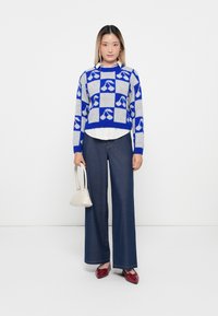 Woman wearing a blue and gray checkered cherry-patterned sweater, white shirt, wide-leg dark jeans, red shoes, holding a small white handbag.