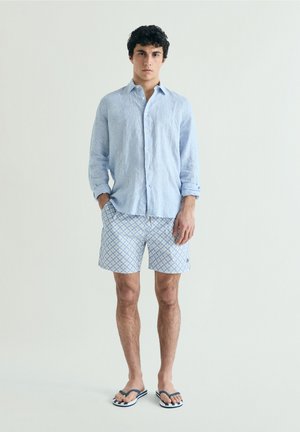 Joven con pelo rizado que lleva una camisa de lino azul claro, pantalones cortos estampados y chanclas azul marino, de pie frente a un fondo liso.