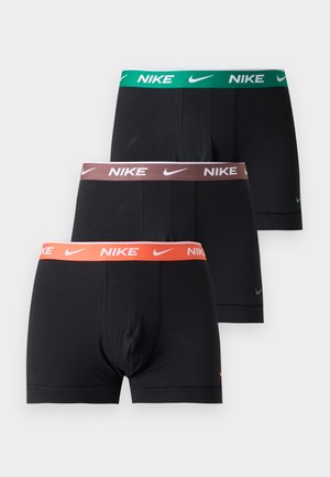 Homme torse nu, debout pieds nus, portant un boxer noir Nike avec une ceinture verte sur un fond blanc uni.