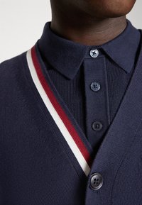 Marinefarbener Cardigan mit rot-weißen Streifenakzenten, geknöpftem Kragen und strukturiertem Strickmaterial. Mit blauen Knöpfen und klassischer Passform.