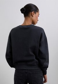 Sudadera negra con cuello redondo, textura suave, hombros caídos y puños de canalé. Combinada con jeans oscuros, vista desde la parte de atrás.