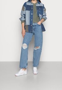 Jeansjacke mit gemischten blauen Patches, kombiniert mit hellblauen, zerrissenen Jeans. Weiße Sneaker mit pinken Akzenten runden das Outfit ab.