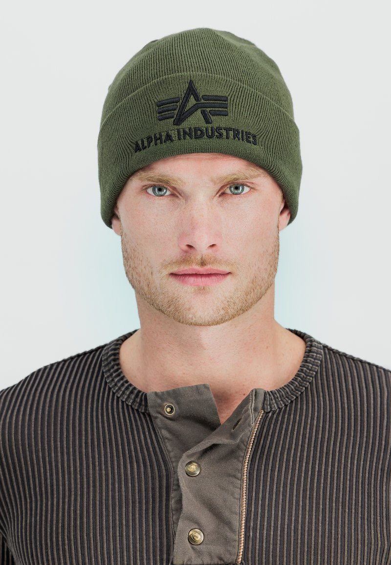 Man with light beard and blue eyes wearing an olive green beanie with "Alpha Industries" logo and a dark ribbed shirt with buttons.