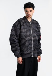 Giacca bomber nera con motivo mimetico, chiusura zip, polsini a coste e tasche laterali. Realizzata in tessuto lucido con una superficie liscia.
