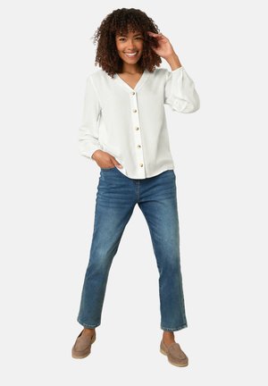 Vrouw met krullend haar, gekleed in een wit blouse met knopen, blauwe spijkerbroek en beige loafers, glimlachend met één hand die haar haar raakt.