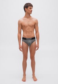 Jeune homme debout pieds nus en slip gris Versace avec ceinture noire, regardant sur le côté, sur un fond clair uni.