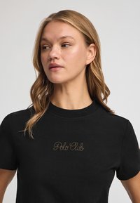 Camiseta de algodón negra con mangas cortas, con texto bordado en dorado "Polo Club" en la parte frontal. Diseño clásico de cuello redondo.