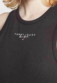 Tumma harmaa hihaton toppi, joka on valmistettu sileästä puuvillasta. Siinä on valkoiset tekstit "TOMMY JEANS New York" ja pieni logo kontrastiväreissä.