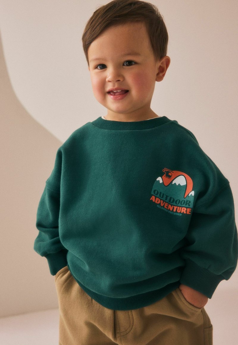 Dark green sweatshirt with a graphic design featuring mountains and "OUTDOOR ADVENTURE" in orange, paired with tan pants.