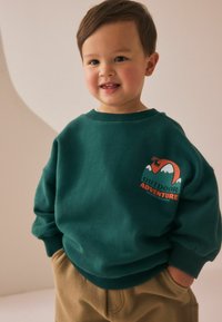 Sweatshirt vert foncé avec un design graphique représentant des montagnes et "AVENTURE EN PLEIN AIR" en orange, associé à un pantalon tan.