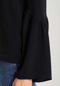 Blusa nera con maniche a campana, realizzata in tessuto leggero. Presenta una superficie testurizzata e dettagli arricciati sulla cucitura della spalla.