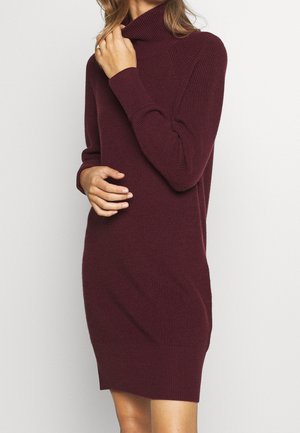 Femme portant une robe pull à col roulé côtelée, manches longues, de couleur bordeaux foncé, touchant le col d'une main et reposant l'autre sur sa taille.