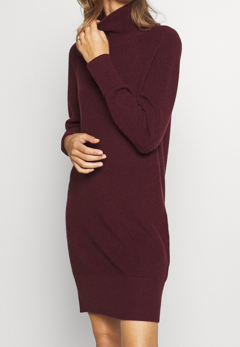 Femme portant une robe pull à col roulé côtelée, manches longues, de couleur bordeaux foncé, touchant le col d'une main et reposant l'autre sur sa taille.