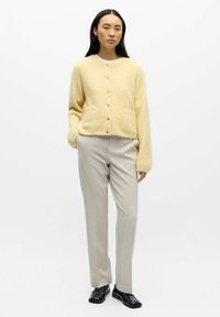 Cárdigan amarillo de punto con cuello redondo, botones delanteros y dos bolsillos, combinado con pantalones entallados beige claro y zapatos de vestir negros.