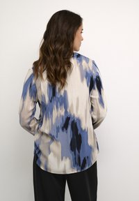 Langärmlige Bluse mit einem abstrakten Muster in Blau, Beige und Schwarz. Hergestellt aus leichtem Stoff, mit abgerundetem Saum und einem Rückendetail.