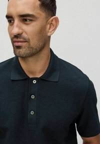 Mörkgrön ribbad polotröja med krage, tre-knapps knäppning och strukturerad tyg. Närbild av övre delen av kroppen och ansiktet.
