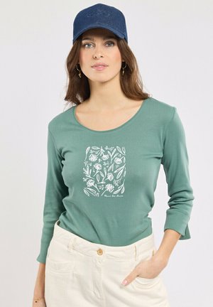 Femme portant une chemise verte à manches longues avec un imprimé floral blanc, un pantalon crème, une casquette bleu marine et des boucles d'oreilles créoles dorées, la main dans la poche.