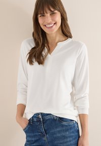 Haut blanc à manches longues avec col et manches trois-quarts, associé à un jean en denim bleu. Le tissu semble lisse et extensible.