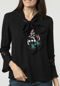 Blusa negra con detalle de cuello atado, que presenta bordados florales coloridos. Manga larga con acentos de encaje en los puños. Textura suave.